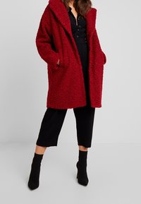 Červená nadměrná bunda s kapucí, chlupatý texturovaný materiál, dvě boční kapsy, nošená přes černý zapínací outfit, doplněná černými vysokými podpatky.