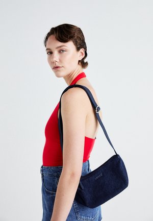 Jonge vrouw met kort bruin haar draagt een rood haltertopje en blauwe spijkerbroek, met een kleine donkerblauwe Levi's schoudertas.