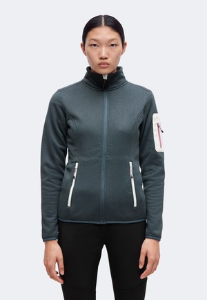 Mujer que lleva una chaqueta con cremallera texturizada de color gris oscuro con bolsillos detallados en blanco y un pequeño bolsillo con cremallera en la manga izquierda.