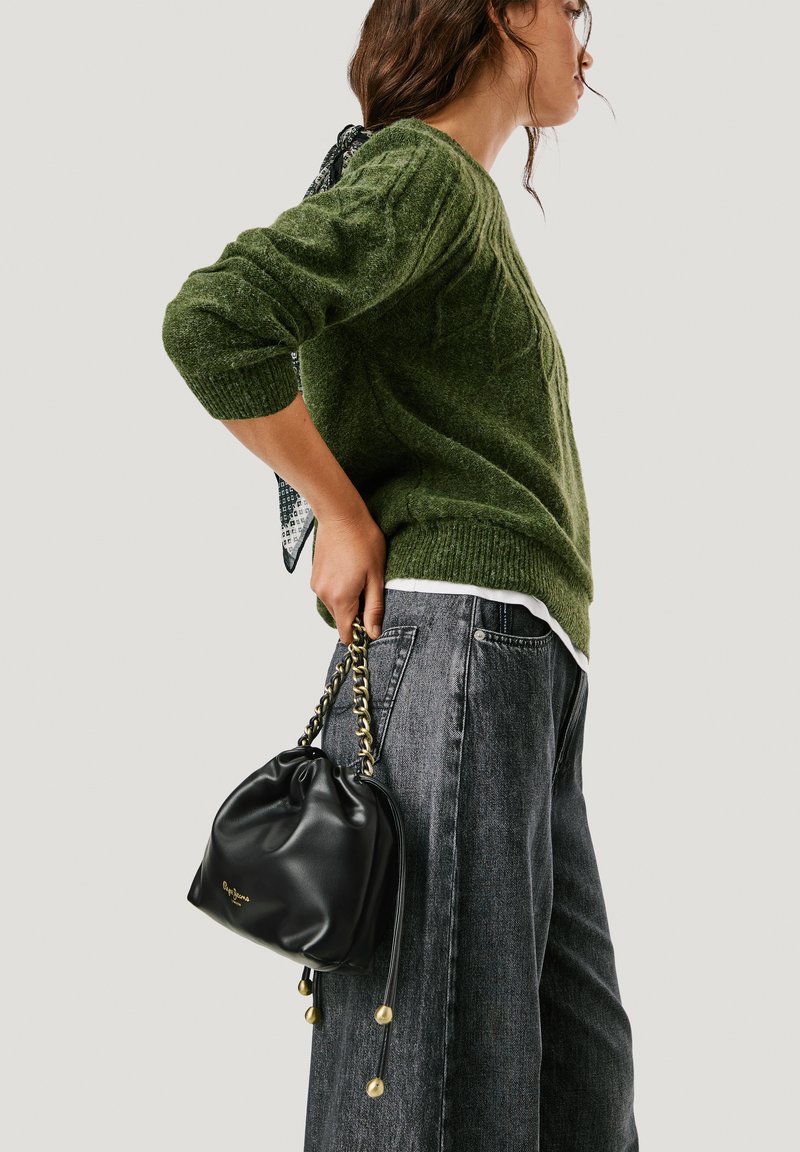 Moteris žalia megztiniu ir pilkomis džinsinėmis kelnėmis laiko juodą odinę rankinę su auksiniu grandinės rankenėle prieš vientisą foną.