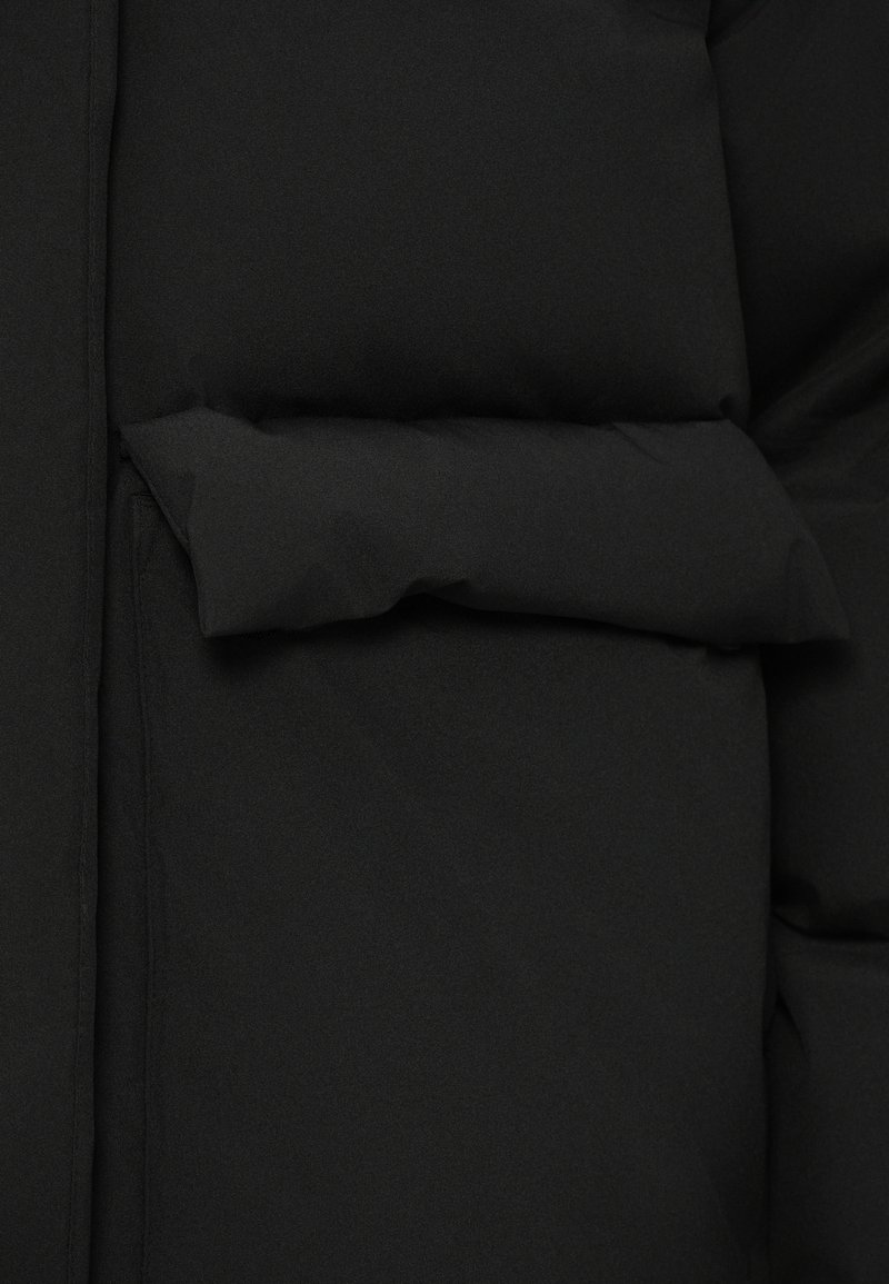 Veste noire matelassée au toucher lisse, avec une couture horizontale prononcée et une poche extérieure intégrée dans le design.