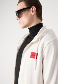 Felpa bianca con zip e un'etichetta rettangolare rossa con la scritta "HUGO," indossata sopra un dolcevita nero. Il modello indossa anche occhiali da sole neri.