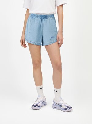 Човек, носещ светлосини дънкови къси панталони Nike, бели чорапи Nike, бели и лилави маратонки Nike и бяла тениска, стоящ на бял фон.