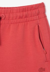 Röda idrottsshorts med elastisk midja, sidoficka med dragkedja och en liten logotypdetalj på tyget. Mjuk, slät textur.