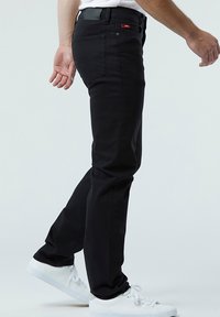 Schwarze Jeans aus Denim mit schlankem Schnitt. Besitzt ein Logo auf der Gesäßtasche, gerade Beine und einen Reißverschluss. Kombiniert mit weißen Sneakers.