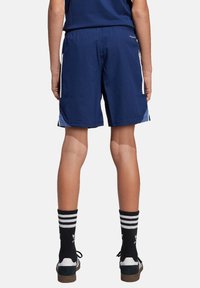 Marinefarbene Sportshorts mit glatter Textur, elastischem Bund und weißen Seitenstreifen. Getragen mit schwarz gestreiften Socken und dunklen Sneakers.