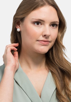 Weiße Perlenohrringe, die von einer Person mit langen, welligen braunen Haaren getragen werden. Das Outfit ist mintgrün und hat eine sanfte Textur.