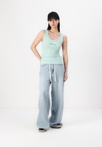Débardeur vert clair avec le logo "GAP", associé à un jean bleu clair à coupe ample et une casquette noire. Les chaussures sont noires et beige, avec une texture mate.