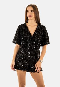 Mono corto de lentejuelas negras con un escote pronunciado en V, mangas cortas y sueltas, y una cintura ajustada. La textura es brillante y refleja la luz.