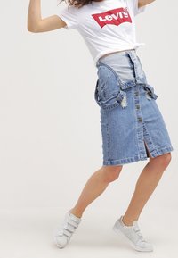 Mujer vistiendo una camiseta blanca de Levi's, una falda de mezclilla azul con botones y tirantes, y zapatillas blancas, posando frente a un fondo liso.