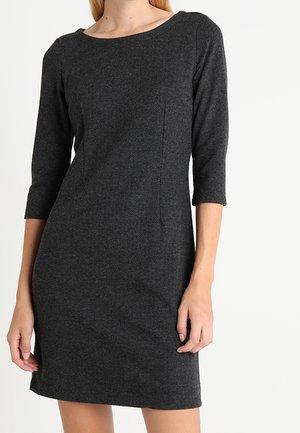 Femme portant une robe cintrée gris foncé avec des manches trois-quarts et un col rond, debout devant un fond clair uni.