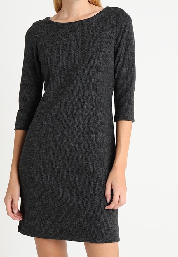 Femme portant une robe cintrée gris foncé avec des manches trois-quarts et un col rond, debout devant un fond clair uni.
