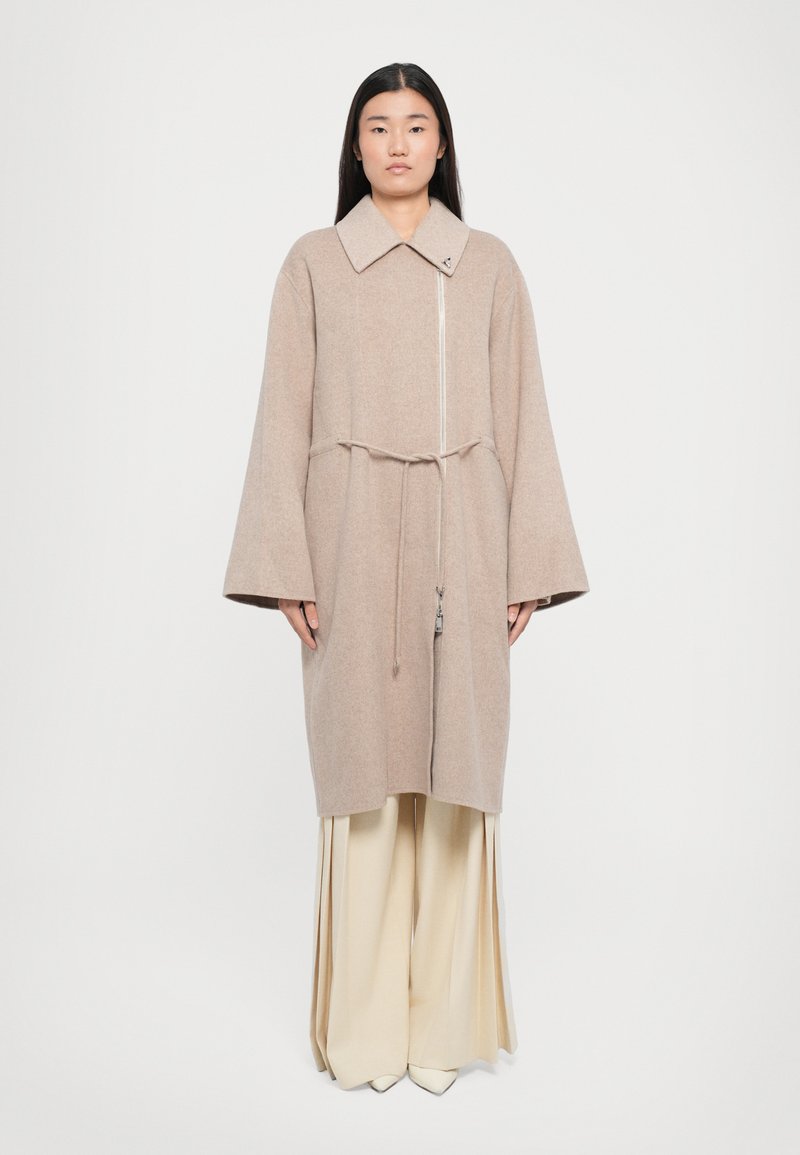 Beige wool blend coat with a large collar, front zip, cinched waist, and wide sleeves. Worn over cream wide-legged trousers.