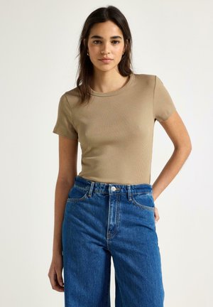 Ung kvinde iført beige kortærmet ribstrikket top og højtaljede blå jeans, stående med den ene hånd på hoften mod en ensfarvet baggrund.