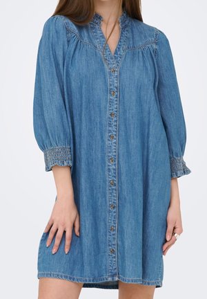 Frau trägt ein blaues Jeanshemdkleid mit Rüschenkragen und gerafften Dreiviertelärmeln, steht mit einer Hand auf dem Kleid abgestützt.