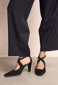 Czarne zamszowe buty na wysokim obcasie z zaostrzonym noskiem i skrzyżowanymi paskami wokół kostki, z błyszczącym obcasem i odsłoniętymi bokami.