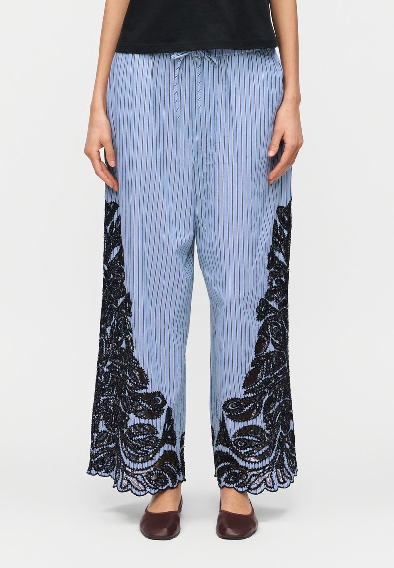 Μπλε ριγέ παντελόνια φαρδιάς γραμμής με μαύρα δαντελένια λουλουδένια σχέδια στα πλάγια και στο κάτω μέρος, φορεμένα με σκούρα καφέ παπούτσια slip-on.