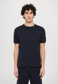 T-shirt azul marinho feito de algodão suave. Decote arredondado, mangas curtas, punhos ligeiramente enrolados e detalhes contrastantes nos ombros. Marca na bainha.