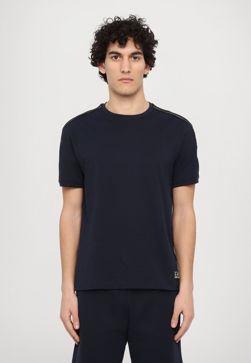 T-shirt azul marinho feito de algodão suave. Decote arredondado, mangas curtas, punhos ligeiramente enrolados e detalhes contrastantes nos ombros. Marca na bainha.