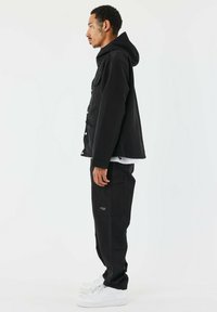 Giacca nera con cappuccio e chiusura a bottoni, tasche laterali, abbinata a pantaloni neri con tasche cargo e sneakers bianche.