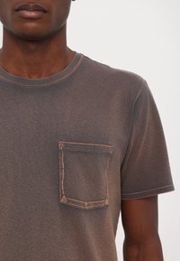Buste masculin portant une chemise marron délavée à manches courtes avec une poche poitrine avant et des coutures visibles sur un fond uni.