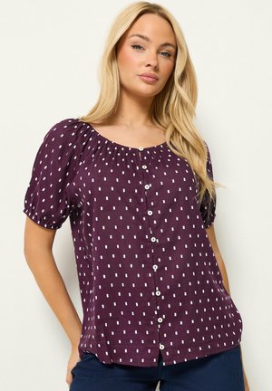 Mujer rubia que lleva una blusa morada de manga corta con patrones cuadrados blancos y botones al frente, combinada con pantalones azul oscuro.