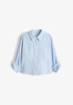 Camicia azzurro chiaro a maniche lunghe con bottoni frontali, due tasche sul petto e maniche arrotolate su sfondo bianco.