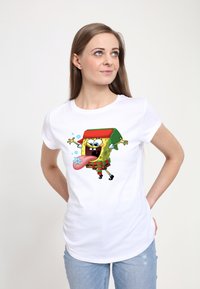 Hvid T-shirt med en farverig tegneseriefigur iført en rød hat, som stikker tungen ud, sat mod en ensfarvet baggrund.