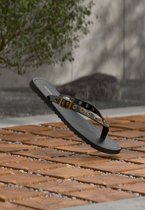 Μαύρο σανδάλι φλιπ φλοπ με χρυσή πόρπη και οπές να αιωρείται πάνω από ξύλινο πλακάκι σε υπαίθριο δάπεδο με γκρι χαλίκι ανάμεσα.