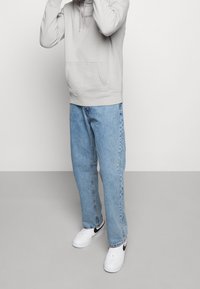 Sudadera con capucha de color gris claro con bolsillo frontal y detalle de cordón, combinada con vaqueros azules holgados y zapatillas blancas con detalles en negro.