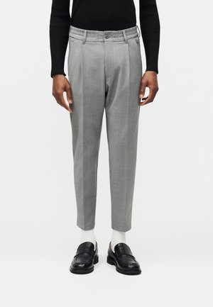 Hombre que lleva pantalones de vestir ajustados de color gris claro, calcetines blancos, mocasines de cuero negro y una camiseta negra de manga larga, de pie sobre un fondo blanco.