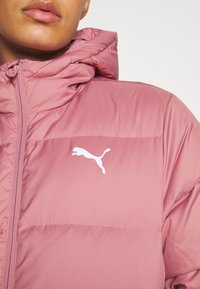 Kurtka z kapturem marki Puma w jasnoróżowym kolorze, z zapięciem na zamek i białym logo na klatce piersiowej. Gładka tekstura i puszysty fason.