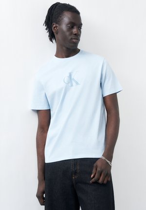 Homme avec des dreadlocks portant un t-shirt CK bleu clair et un jean foncé, regardant vers sa gauche sur un fond clair uni.