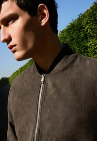 Chaqueta bomber de ante marrón con cuello de canalé, que presenta una cremallera plateada. La textura es suave, sin patrones ni acentos visibles.