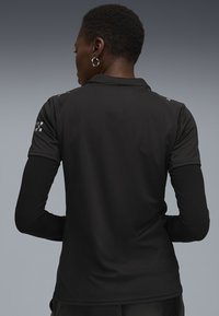 Person med kort hår iført en sort polo-shirt over en sort langærmet bluse, der står med ryggen til en grå gradient baggrund.