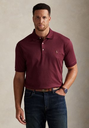 Mand iført en mørkerød polo med lille logo og mørkeblå jeans, stående med den ene hånd i lommen mod en ensfarvet baggrund.