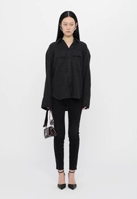 Black long-sleeve button-up shirt with two chest pockets, paired with slim black pants and pointy black heels. Holding a small shiny handbag.