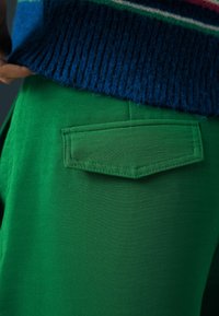 Groene broek van een textuurstof met een rechthoekige achterzak. Een blauwe, geribbelde trui is gedeeltelijk zichtbaar boven de tailleband.
