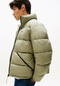 Chaqueta de plumas verde oliva con cuello alto, textura acolchada, detalles en cremallera negra y un logo en el pecho izquierdo.