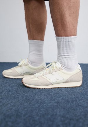 Osoba w kremowych sneakersach New Balance i białych skarpetkach stoi na niebieskim dywanie; widoczne są tylko dolne partie nóg i buty.