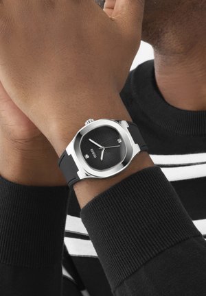 Schwarze Guess-Armbanduhr mit Lederarmband, silbernem quadratischem Gehäuse und schwarzem strukturiertem Zifferblatt, getragen am linken Handgelenk vor einem schwarz-weiß gestreiften Pullover.