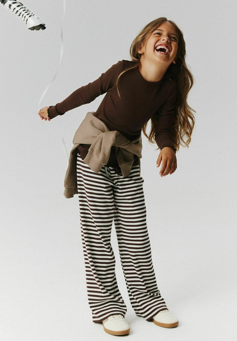 Jeune fille riant et se penchant en avant tenant une ficelle de ballon, portant un t-shirt marron à manches longues, un pantalon rayé et des chaussures blanches.