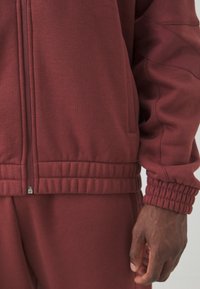Bordo zip-up jakna s rebrastim manžetama i rubom, izrađena od mekanog materijala. Ima bočne džepove i blago strukturiranu površinu.