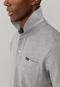 Man draagt een lichtgrijs poloshirt met omhoogstaande gestreepte kraag en een borstzak met een klein donkerblauw label.