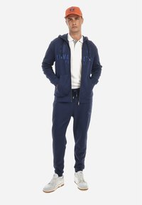 Navy modra zip-up jopica in hlače s logotipom, v kombinaciji z belo polo majico in oranžno kapo. Bele športne čevlje z teksturiranimi podplati.