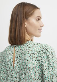 Patroonblouse met groene en teal bladeren op een witte achtergrond. De achterkant heeft een sleutelgatopening met een knoopsluiting. Gelaagde mouwen.