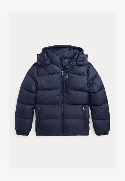 Veste matelassée bleu marine avec fermeture éclair devant, trois poches zippées, capuche amovible, poignets élastiques et logo rouge brodé sur la poitrine gauche.