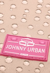 Etiqueta de goma rosa con el texto "JOHNNY URBAN JUNIOR" sobre un fondo beige texturizado, con gotas de agua, indicando resistencia al agua.