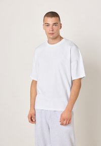 Bela bombažna majica s kratkimi rokavi z okroglim izrezom, sproščenim krojem in gladko teksturo, kombinirana s sivimi trenirkami.