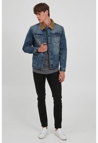 Denim jacket with a brown fur collar, faded blue color, buttoned front, two chest pockets, worn over a gray t-shirt and black pants.